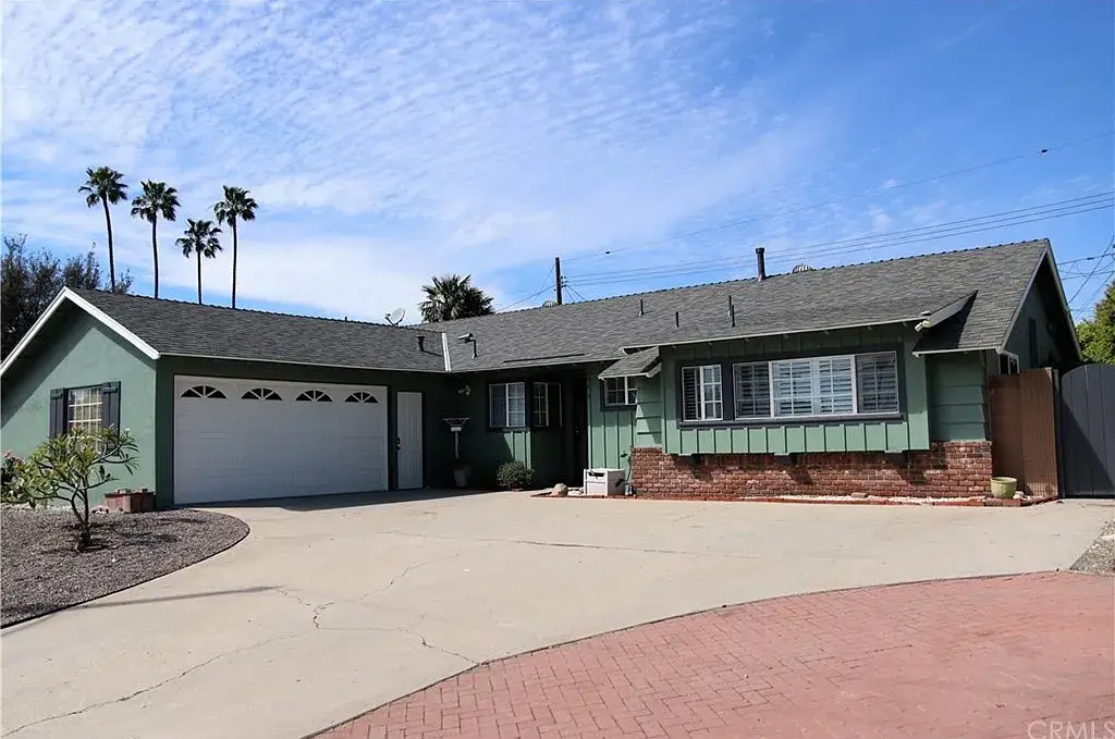 Green single-story house with double garage, brick accents, palm trees, and driveway.