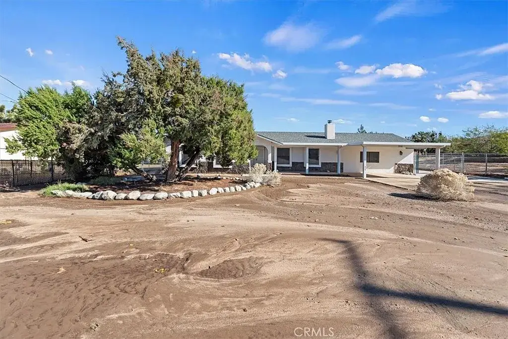 Single-story house with wide porch, sandy yard, sparse trees, and clear blue sky.