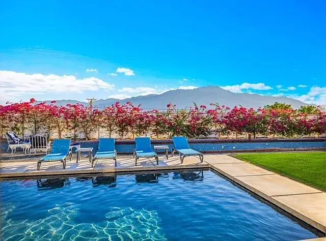 Poolside patio with blue lounge chairs, pink flowers, and mountain views under a clear sky.