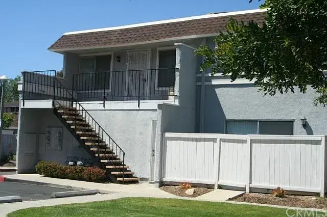 Two-story gray building with brown roof, outdoor staircase, white fence, and greenery.