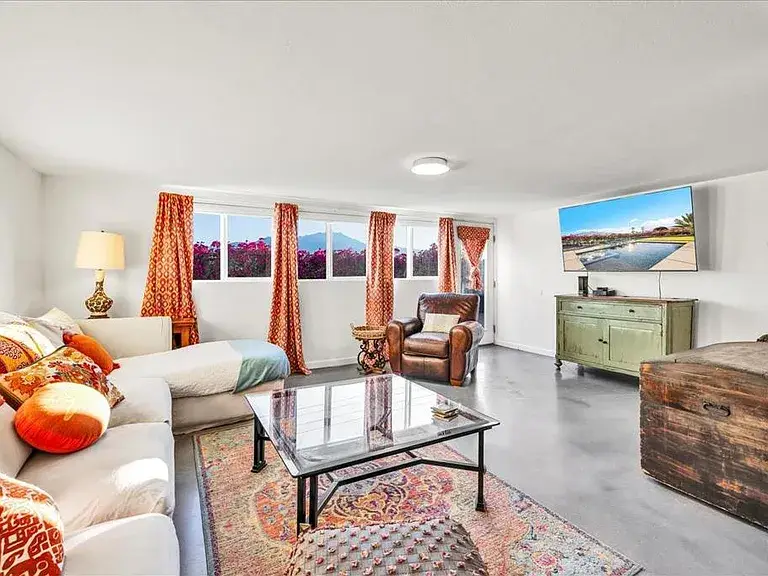 Bright living room with cozy seating, patterned rug, orange accents, and large windows.